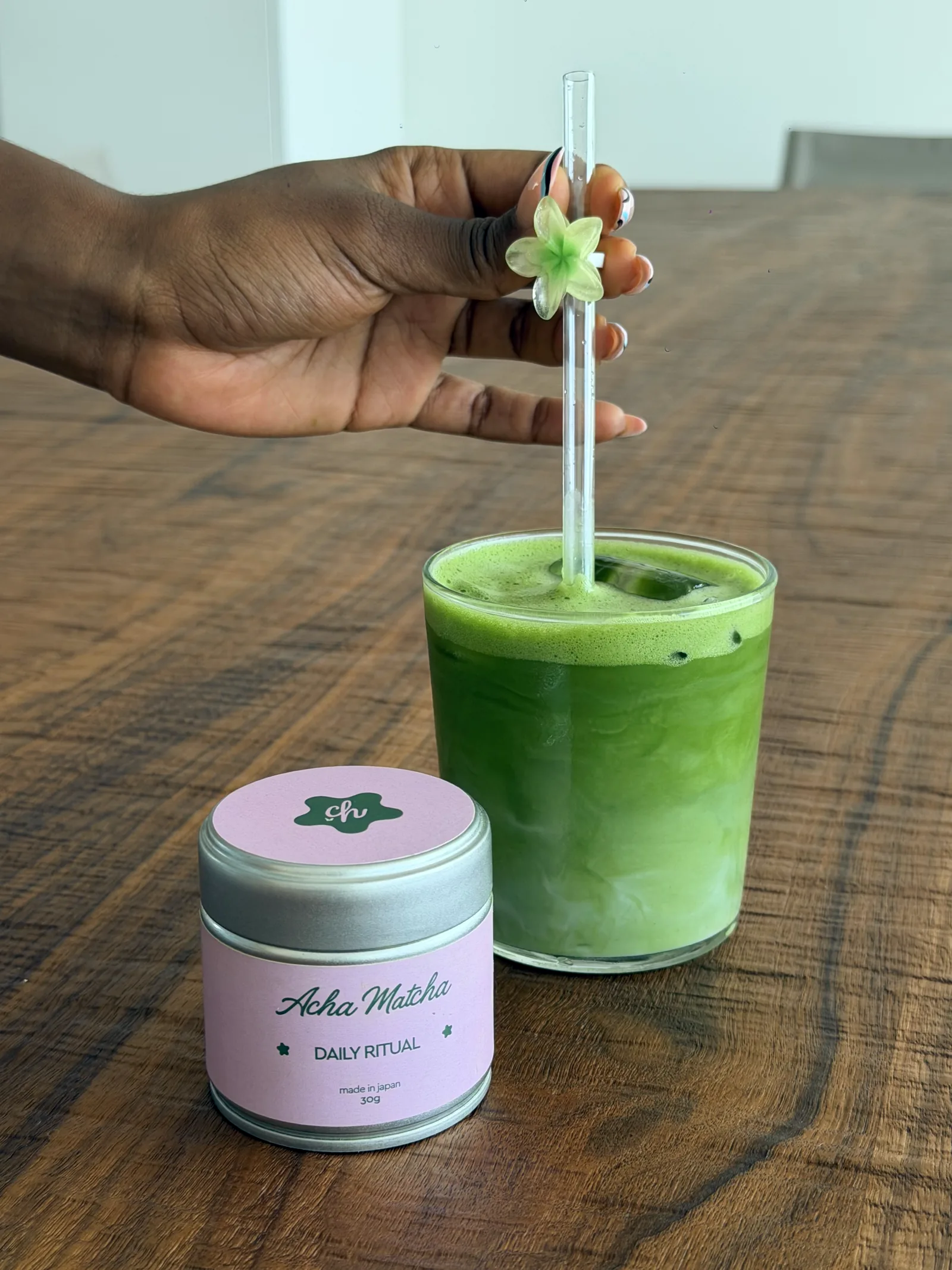 A hand lowering a flower straw into an iced matcha on a walnut table