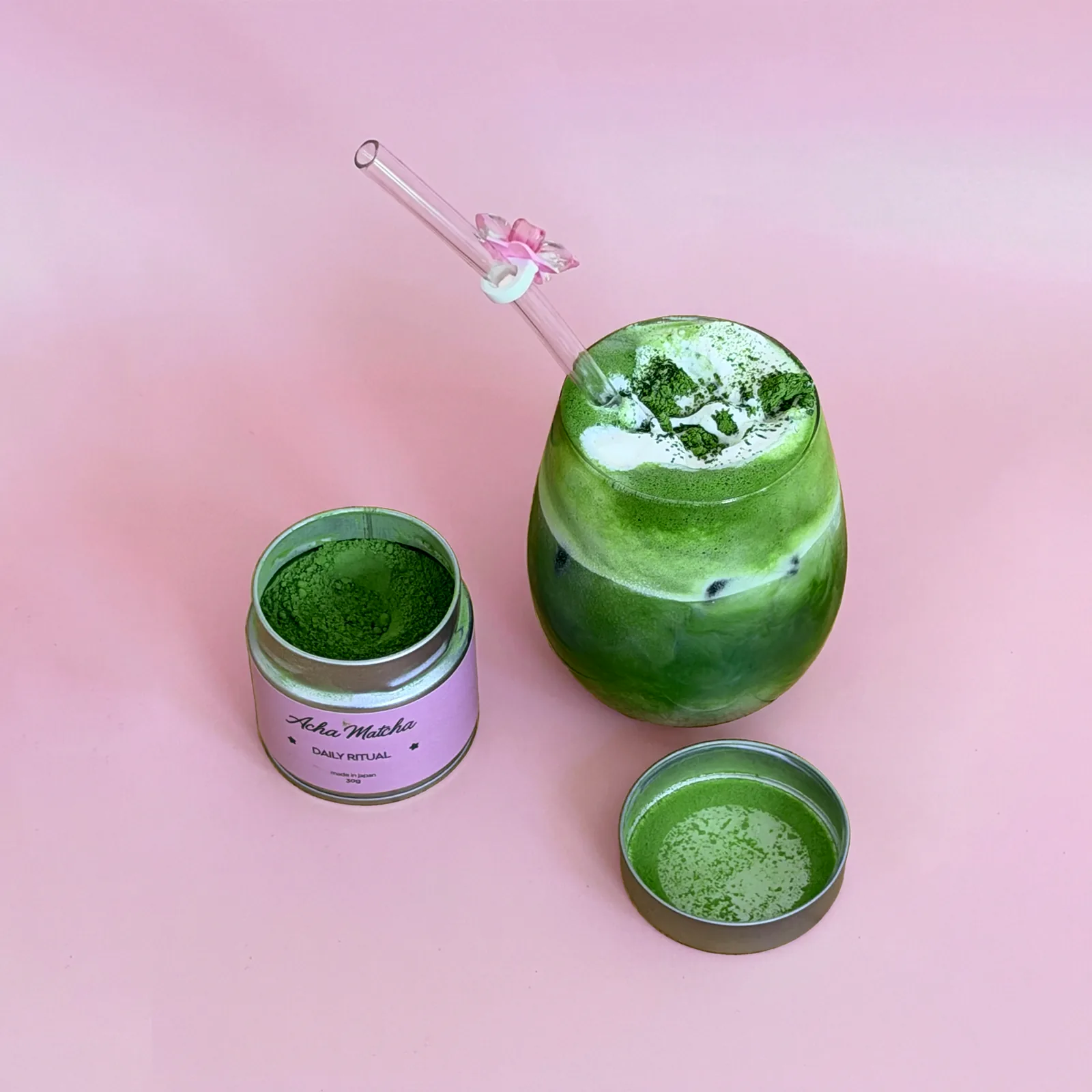 Acha Matcha tin open beside an iced matcha, shot from above on pink