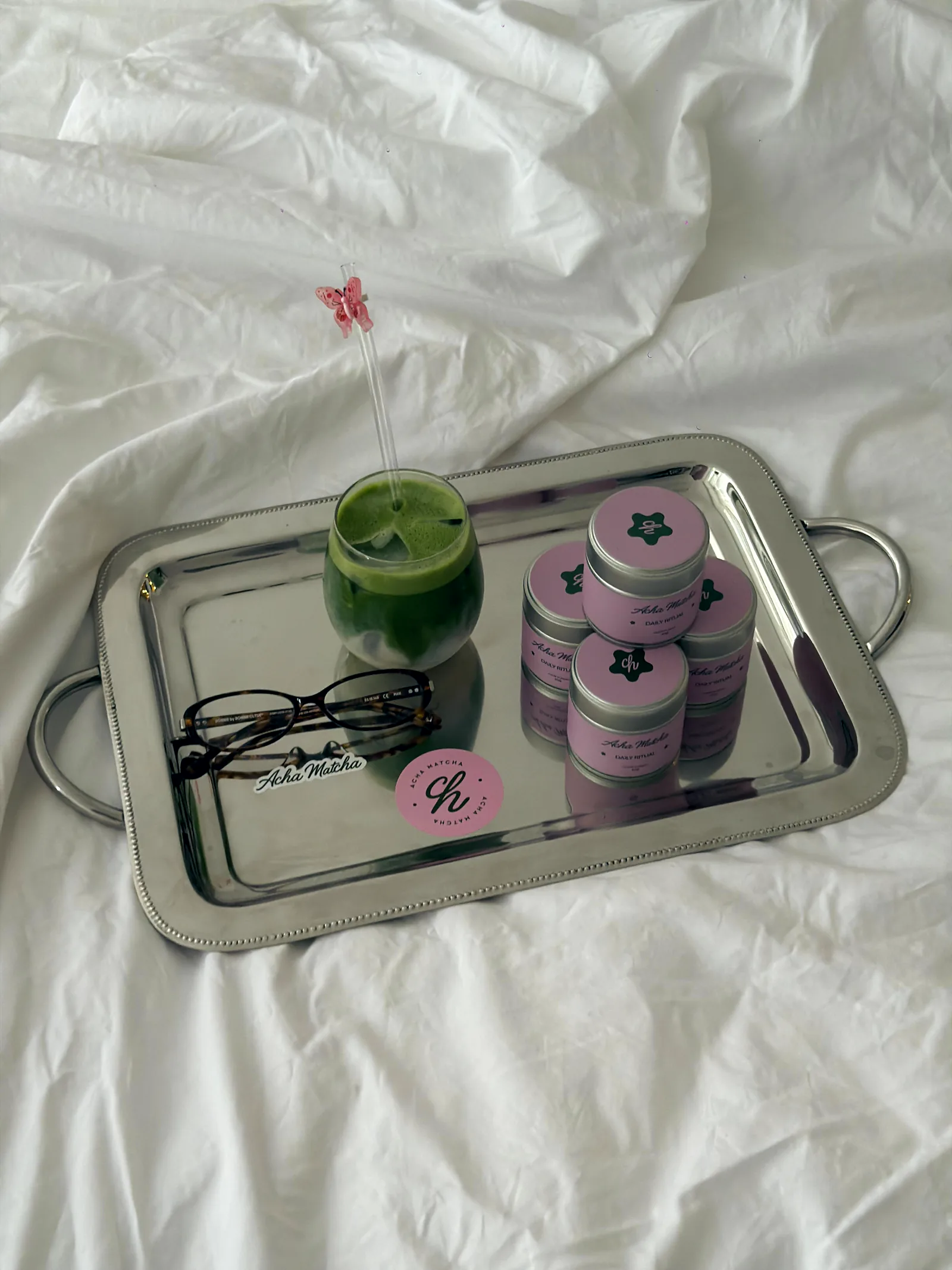 A silver tray of Acha Matcha tins and an iced matcha on white bedding